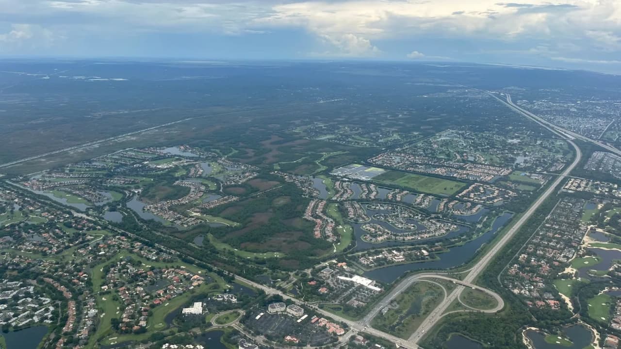 High-altitude aerial over Palm Beach County, Florida, looking across the western edge of suburban development where gated communities and golf courses give way to open wetland, with a turnpike corridor running north to south.